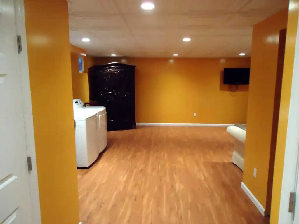 A basement room with orange-yellow walls, wood-like flooring, a washing machine and dryer, a dark armoire, wall-mounted TV, and a light couch—perfect inspiration for home improvements in Nassau County, NY. Recessed ceiling lights brighten the space.