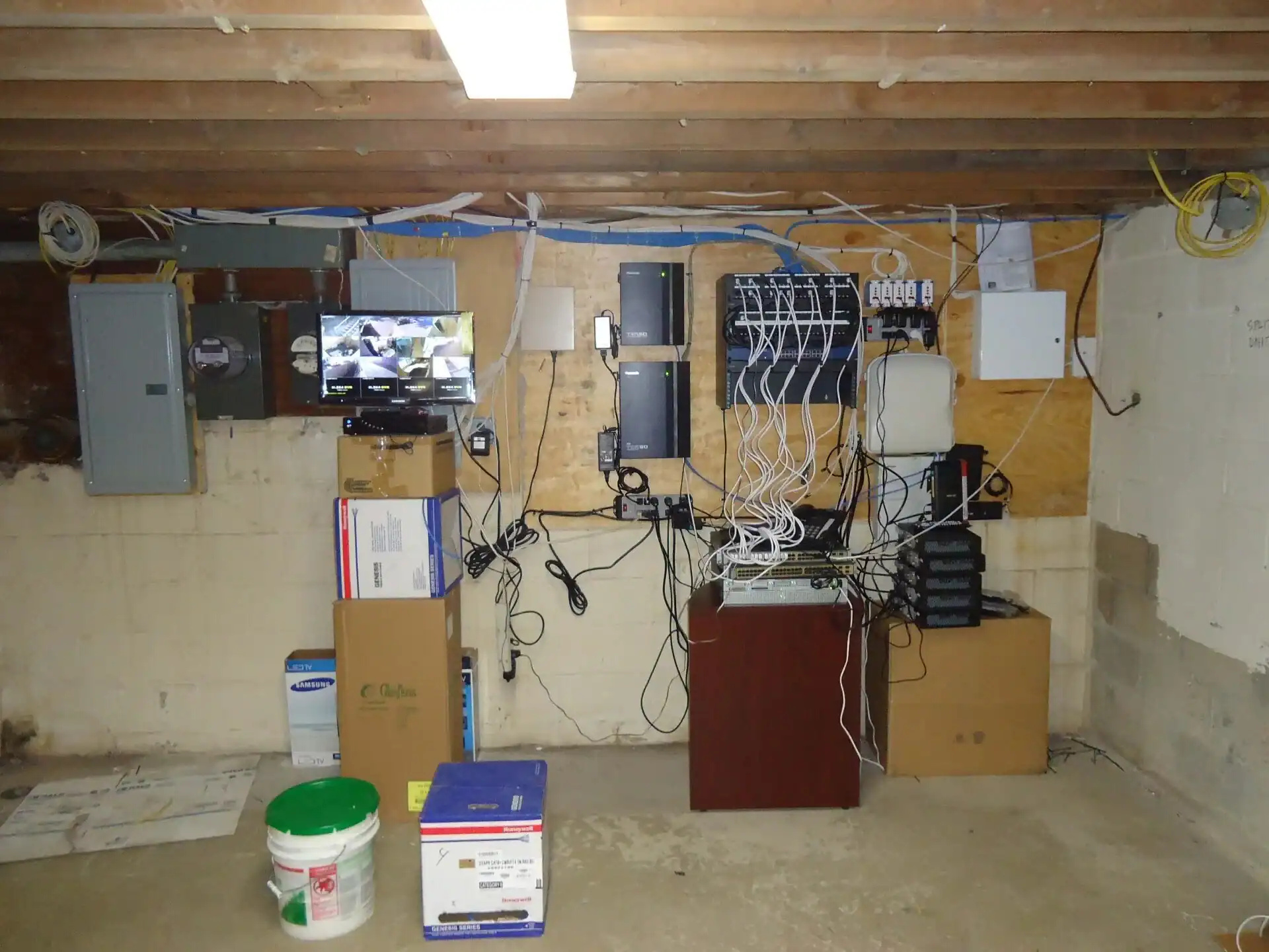 A basement scene in Nassau County, NY highlights electrical panels, surveillance monitors, and network equipment mounted on a wall for recent home improvements. Cardboard boxes and a white bucket rest on the concrete floor beneath exposed ceiling beams.