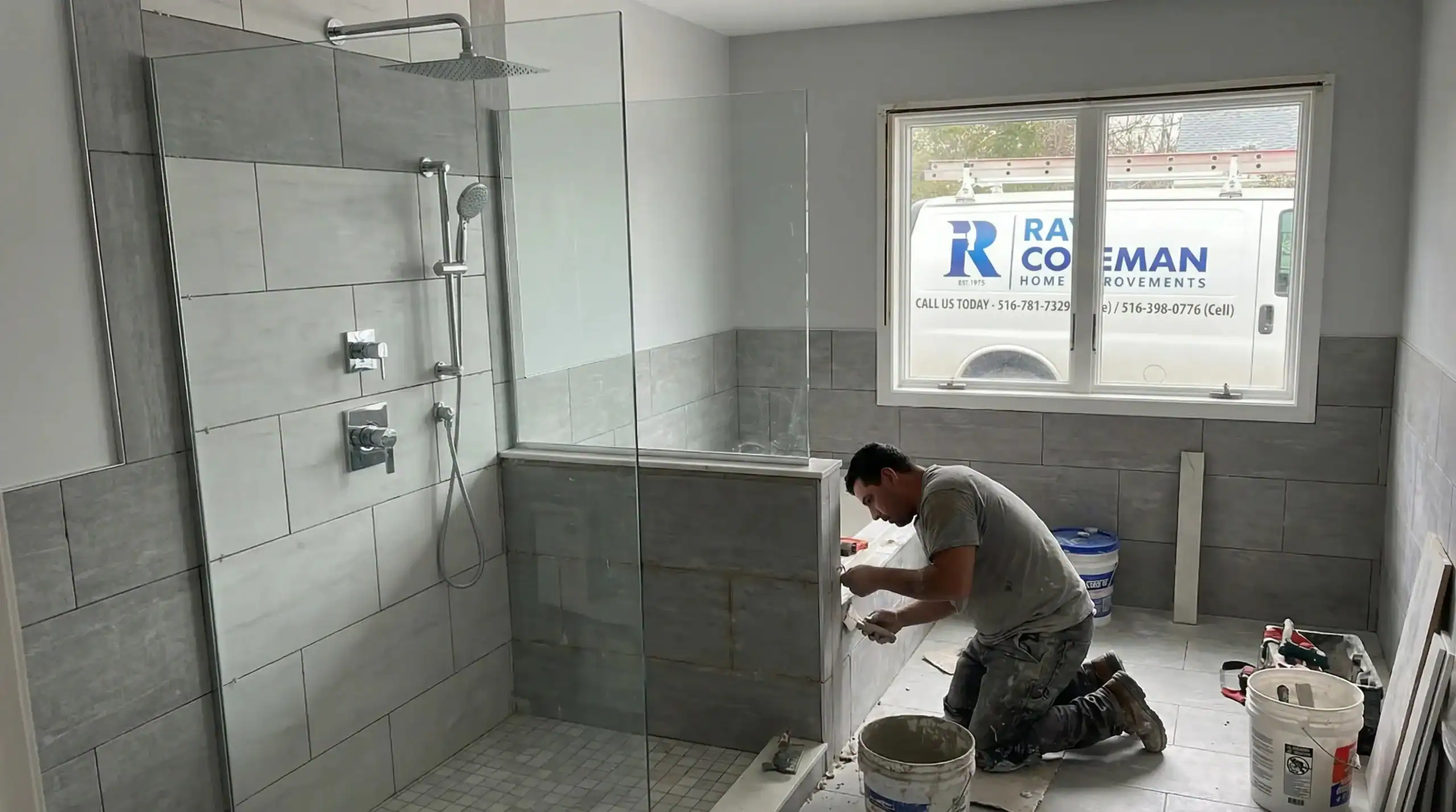 A man kneels and works on tiling in a modern NY bathroom under renovation, with gray tiles and a glass shower enclosure. Daylight enters through a window, where a home improvements Nassau County contractor’s van is visible outside.