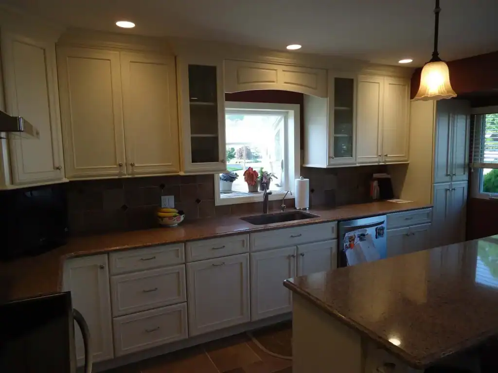 A modern kitchen with cream-colored cabinets, brown countertops, a central sink under a window, and a fruit bowl on the counter—perfect inspiration for home improvements in Nassau County, NY. Recessed ceiling lights and a hanging lamp illuminate the kitchen island.