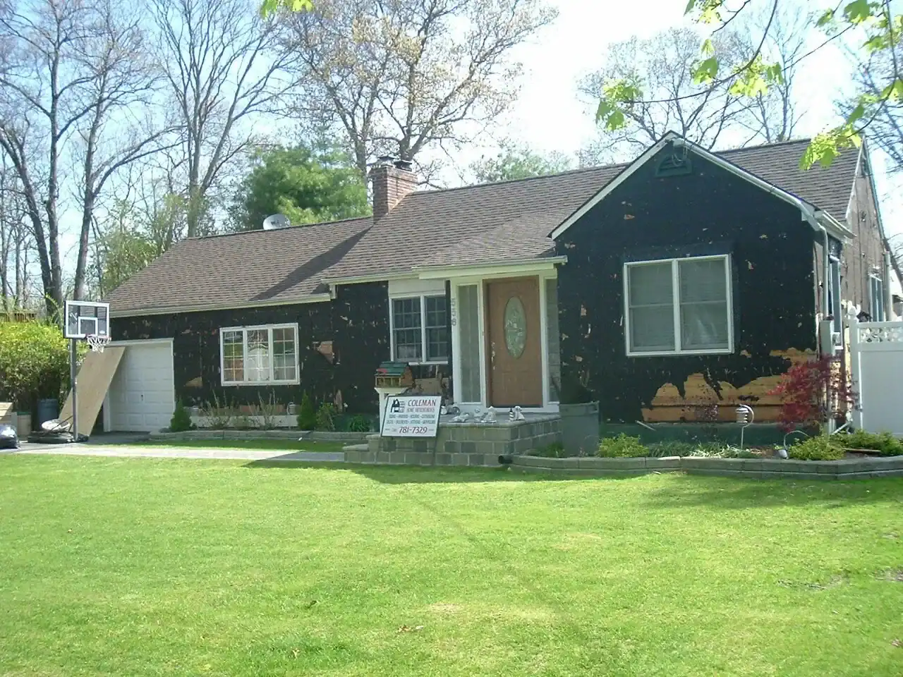 A single-story NY house with dark siding partially removed, showing patches of exposed wood. Home improvements Nassau County materials and a contractor sign are in the yard. The green lawn is well-kept.