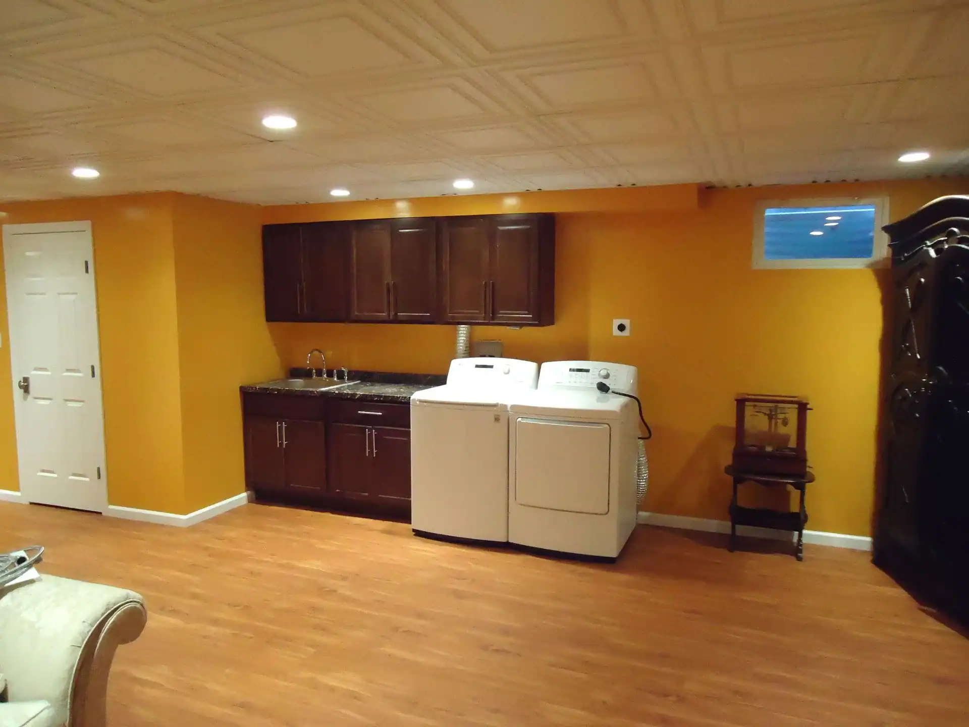 A laundry area in a NY basement with yellow walls, wood flooring, washer and dryer, dark wood cabinets with a sink, a small window, and a side table beside a large armoire—perfect for home improvements Nassau County residents enjoy.
