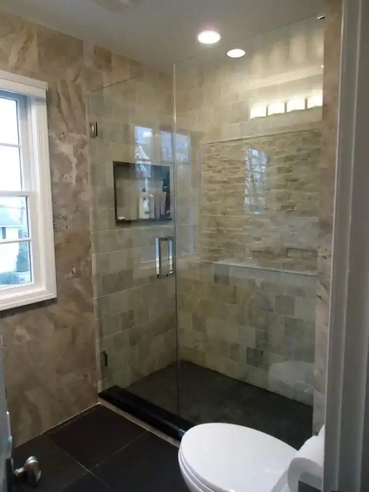 Modern NY bathroom with a glass-enclosed shower featuring beige stone tiles, a built-in shelf, and a window to the left. Perfect for home improvements Nassau County; a portion of a white toilet is visible in the foreground.