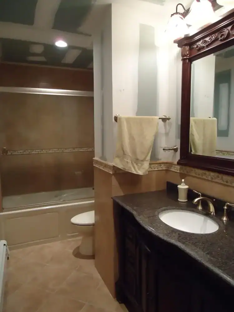 A bathroom with unfinished drywall—a perfect candidate for home improvements in Nassau County, NY—features a bathtub with glass doors, a toilet, dark wood vanity with sink, large mirror, beige towels on racks, and a soap dispenser on the counter.