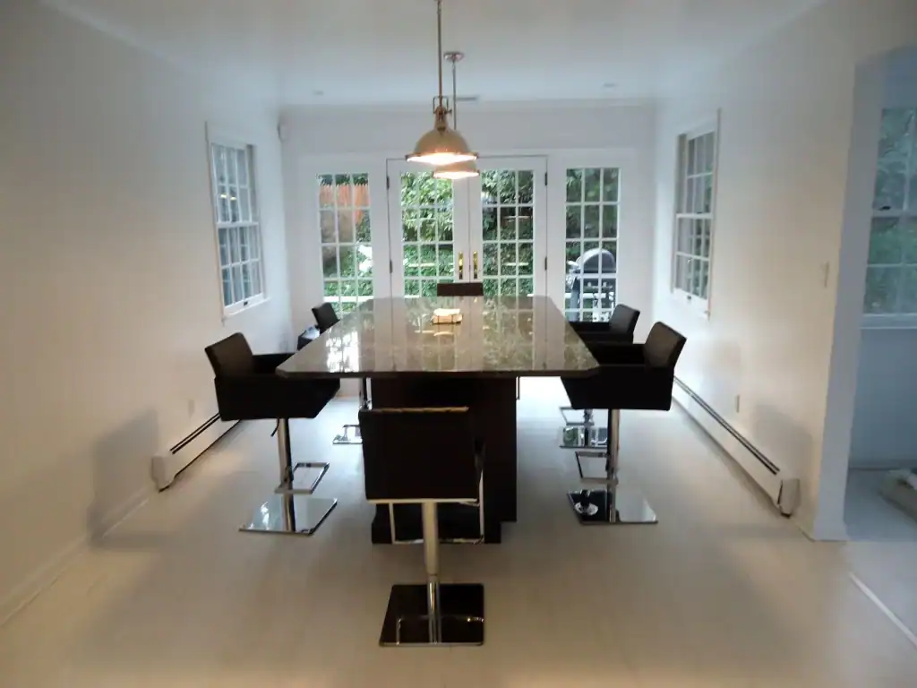 A modern NY dining room with a marble-topped rectangular table, six black high-back chairs, white walls and floors, large windows, and glass doors facing a green garden—ideal for stylish home improvements in Nassau County.