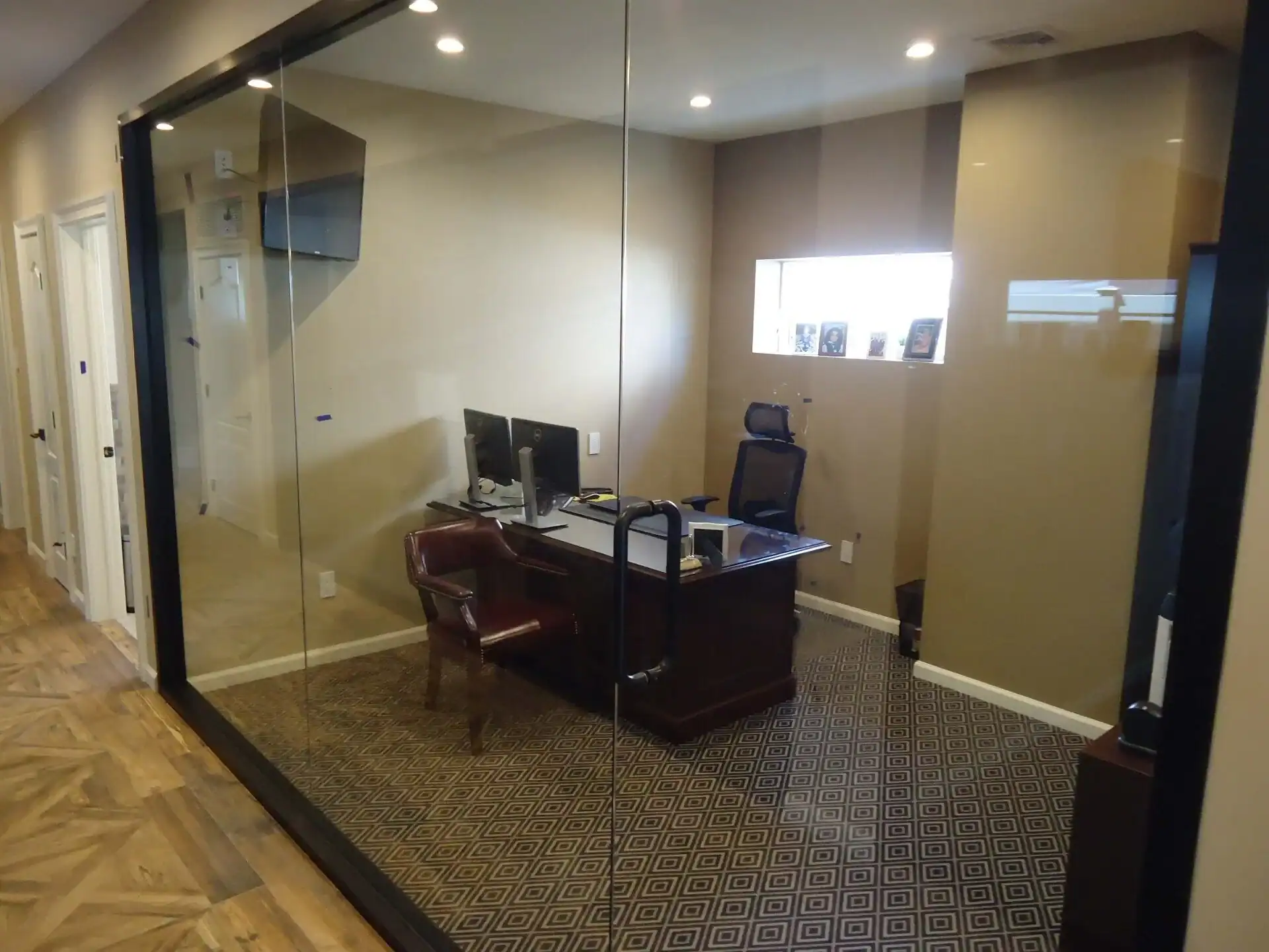 A modern office with glass walls, featuring a large wooden desk, an office chair, guest chair, monitor, and framed photos on a windowsill—perfect inspiration for home improvements in Nassau County, NY. The space is well-lit with overhead lights.