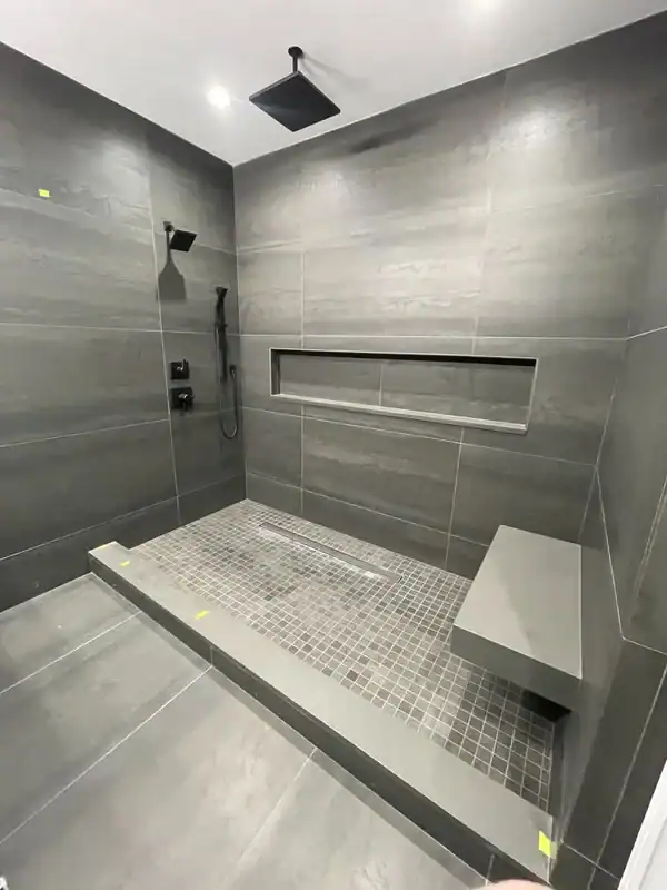 Modern shower with dark gray tiles, a built-in bench, recessed shelf, rainfall showerhead, and handheld shower—perfect for NY home improvements. This minimalistic design features clean lines and a spacious layout ideal for Nassau County homes.