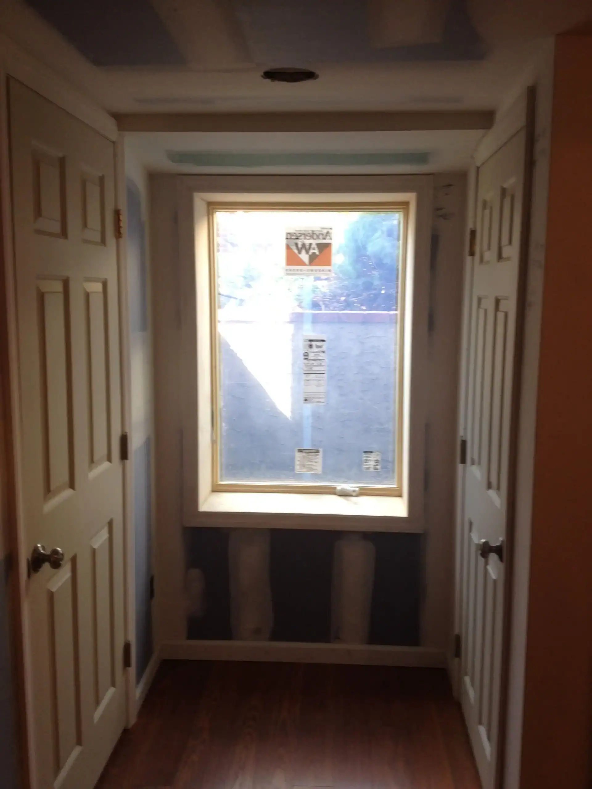 A hallway with wooden floors leads to a window adorned with stickers, flanked by two closed doors on each side. Ideal for those considering home improvements in Nassau County, NY, the window looks out to a blue-lit window well.