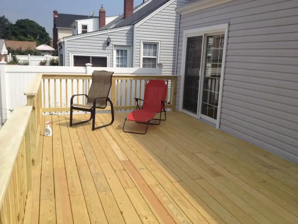 A wooden deck with two chairs—one brown and one red—sits outside a gray house with sliding glass doors and a wooden railing, showcasing stylish home improvements in Nassau County, NY. Nearby houses are visible in the background.