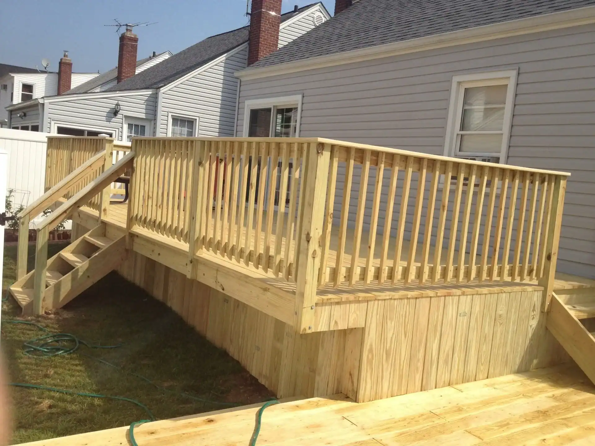 A newly built wooden deck with railings and stairs attached to a light gray NY home. Elevated above the grass, this home improvement in Nassau County features vertical balusters along the railing for added safety and style.